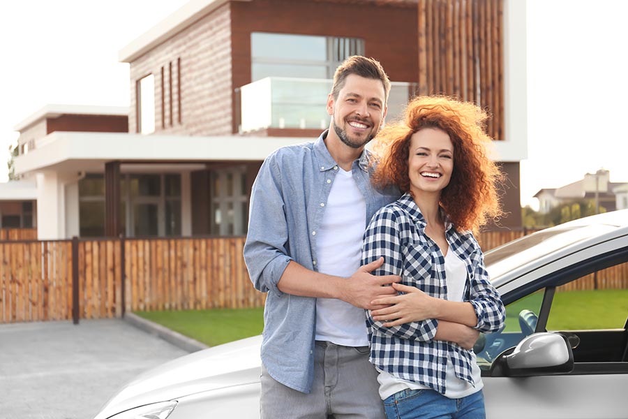 About Our Agency - Couple Pose Outside Their Large Home With Their Silver Car Parked in the Driveway