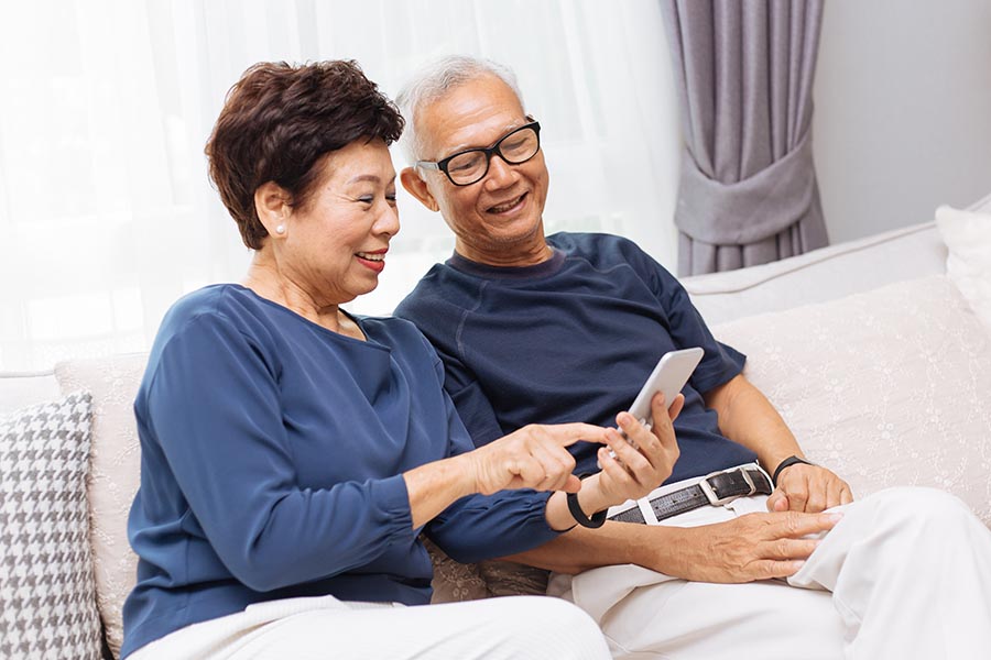 Client Center - Senior Couple Dressed in Blue and White Use a Phone on Their Couch in a Bright Living Room