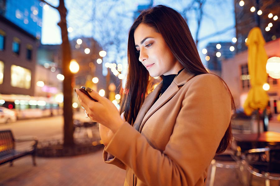 Contact Us - Young Woman in Camel Coat Uses Her Phone on a Sidewalk, as the Businesses Light up Around Her